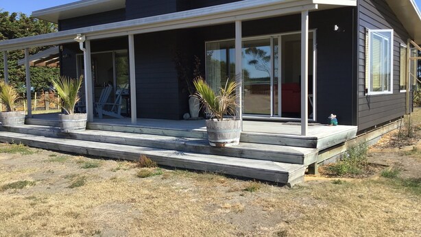 A modern two-story house with a covered porch, large sliding glass doors, and potted plants. - Ready for a cosy Beach holiday? (Foxton Beach)