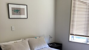 A neatly made bed with white linens, a bedside table with a lamp, a framed picture on the wall, and a window with blinds. - Ready for a cosy Beach holiday? (Foxton Beach)