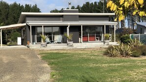 A modern house with a covered porch, large windows, and a well-maintained lawn. - Ready for a cosy Beach holiday? (Foxton Beach)