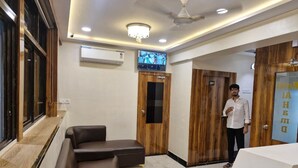 A hotel reception area with a reception desk, a seating area with a sofa and armchairs, wooden doors, and a television mounted on the wall. - Hotel AL-HAMD (Mumbai)