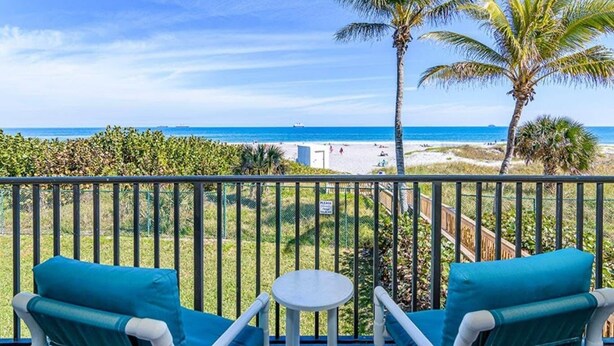 Ein Balkon mit zwei blauen Stühlen und einem kleinen Tisch, mit Blick auf einen Strand mit Palmen und einem strahlend blauen Himmel.