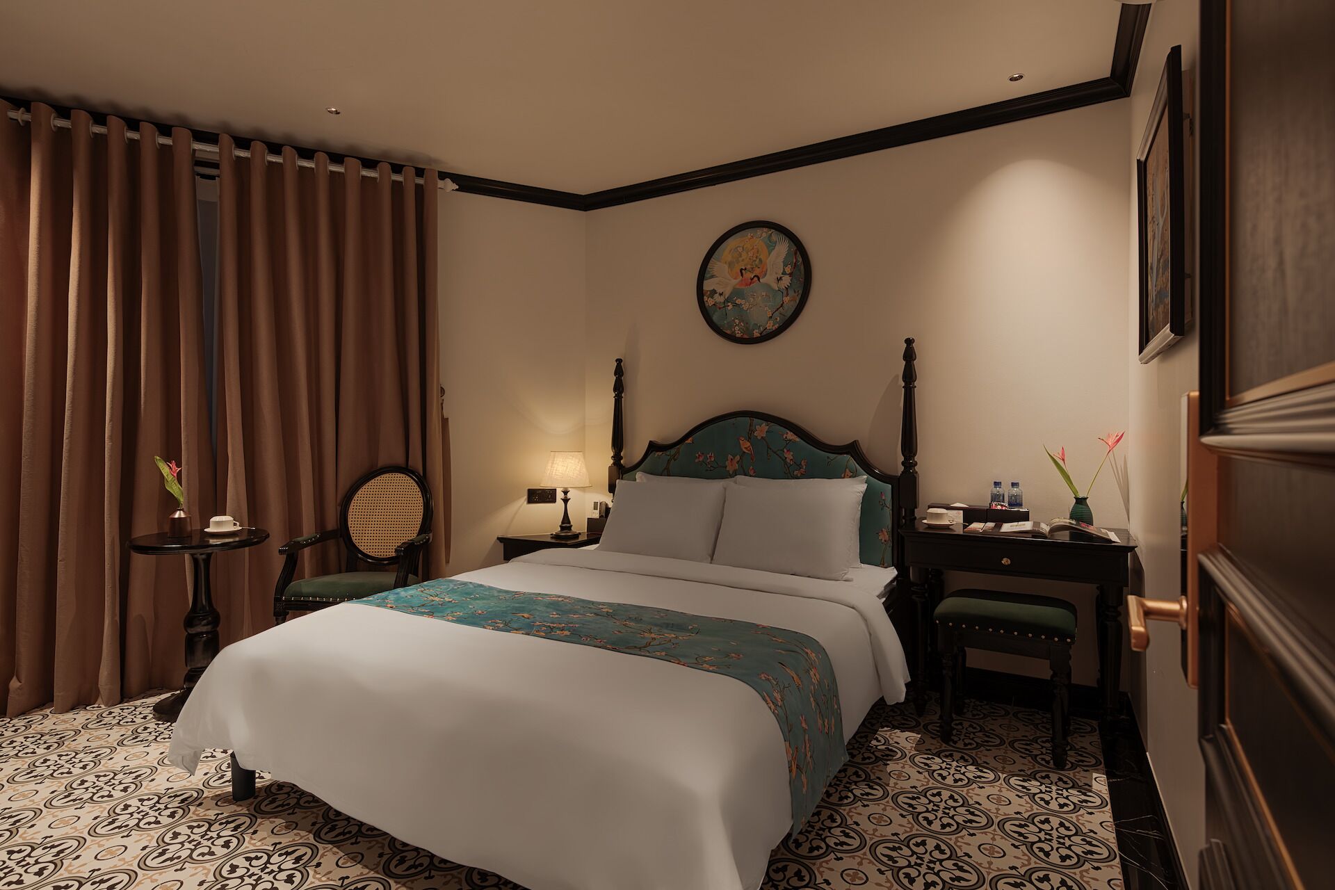A hotel room with a bed, a desk, a chair, a nightstand, a lamp, a vase with flowers, and a painting on the wall.
