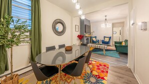 Ein modernes Wohnzimmer mit Essbereich, ausgestattet mit einem runden Glastisch, schwarzen Stühlen, einem farbenfrohen Teppich und einer grünen Pflanze.
