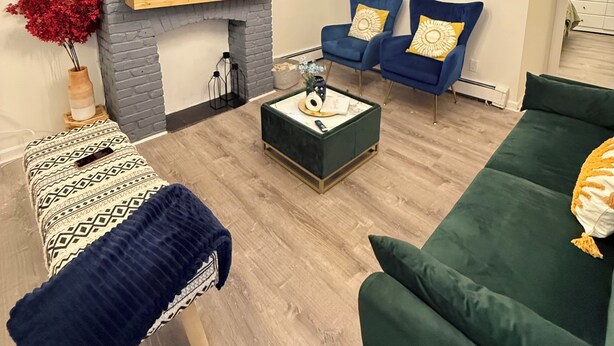 Ein Wohnzimmer mit einem Flachbildfernseher, einem Kamin, einem grünen Sofa, einem blauen Sessel und einem gemusterten Hocker.