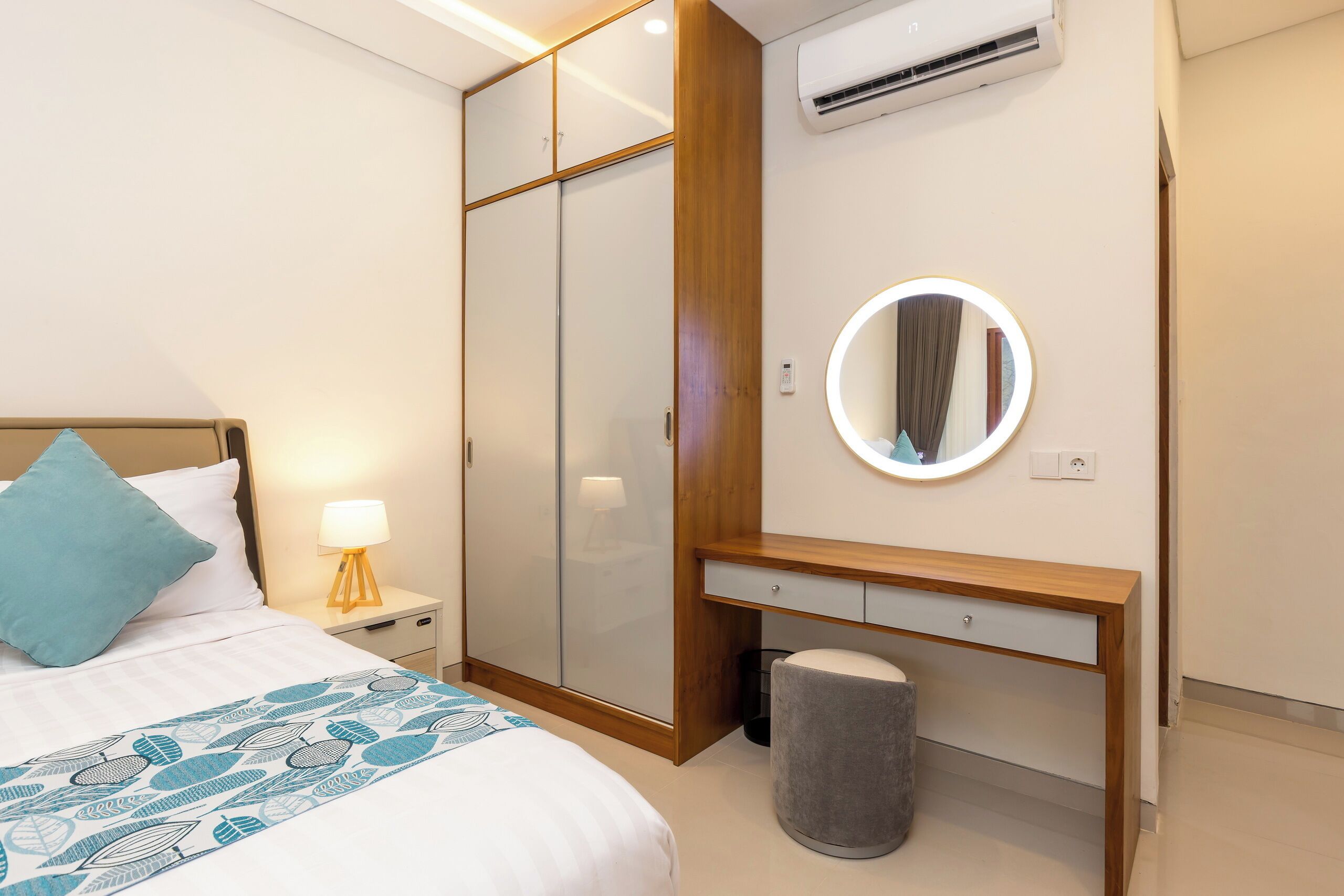 A bedroom with a bed, a bedside table with a lamp, a wardrobe, a vanity with a mirror, and an air conditioning unit.