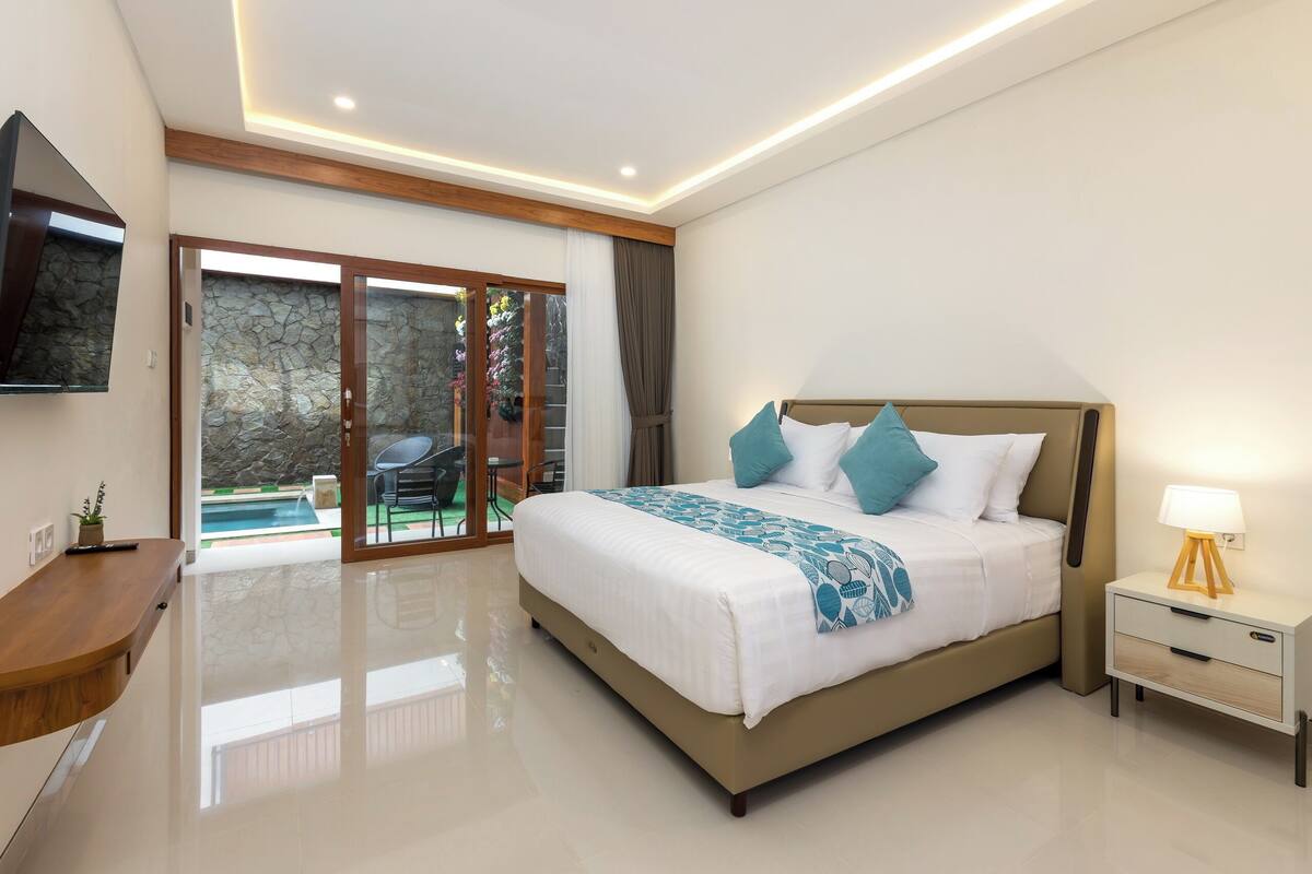A modern bedroom with a large bed, bedside tables, a TV, and a view of an outdoor pool area.