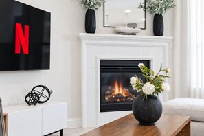 A modern living room with a fireplace, a flat-screen TV mounted above it, and a vase with flowers on a wooden table. - Mid-Stay Relocation | Office, Fast WI-FI Family ready 30+ Stay (Royse City)