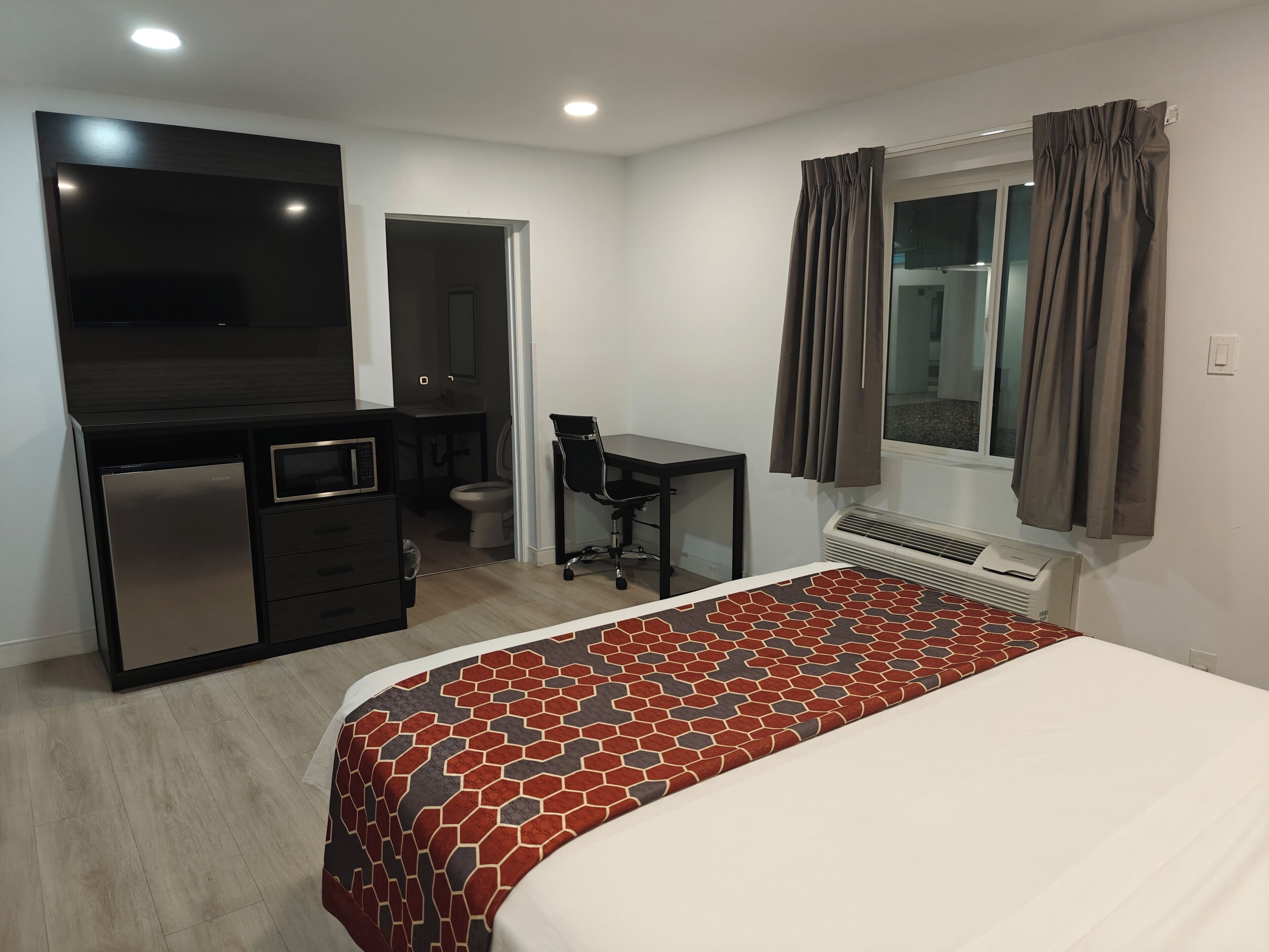 Ein Schlafzimmer mit einem Bett, einem Schreibtisch, einem Stuhl, einem Fernseher, einer Minibar und einem Fenster mit Vorhängen.
