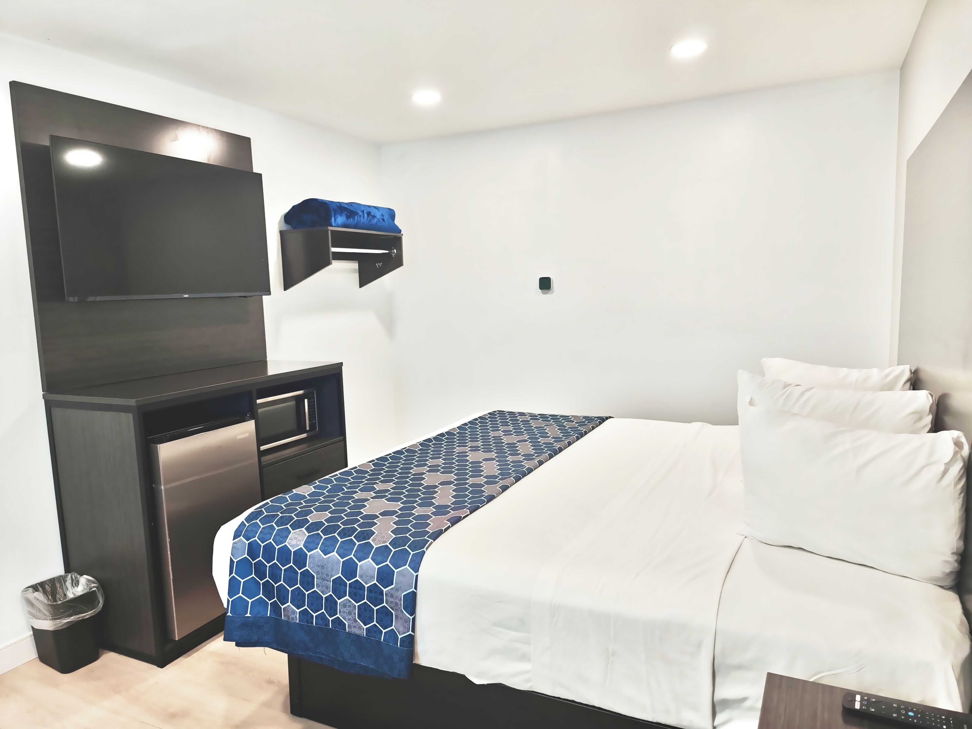 A hotel room with a bed, a TV mounted on the wall, a desk with a computer, and a trash bin.