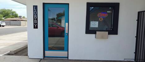 Entrada de un edificio con una puerta azul, un cartel que dice 'LOBBY' y una ventana con el letrero 'ABIERTO'.