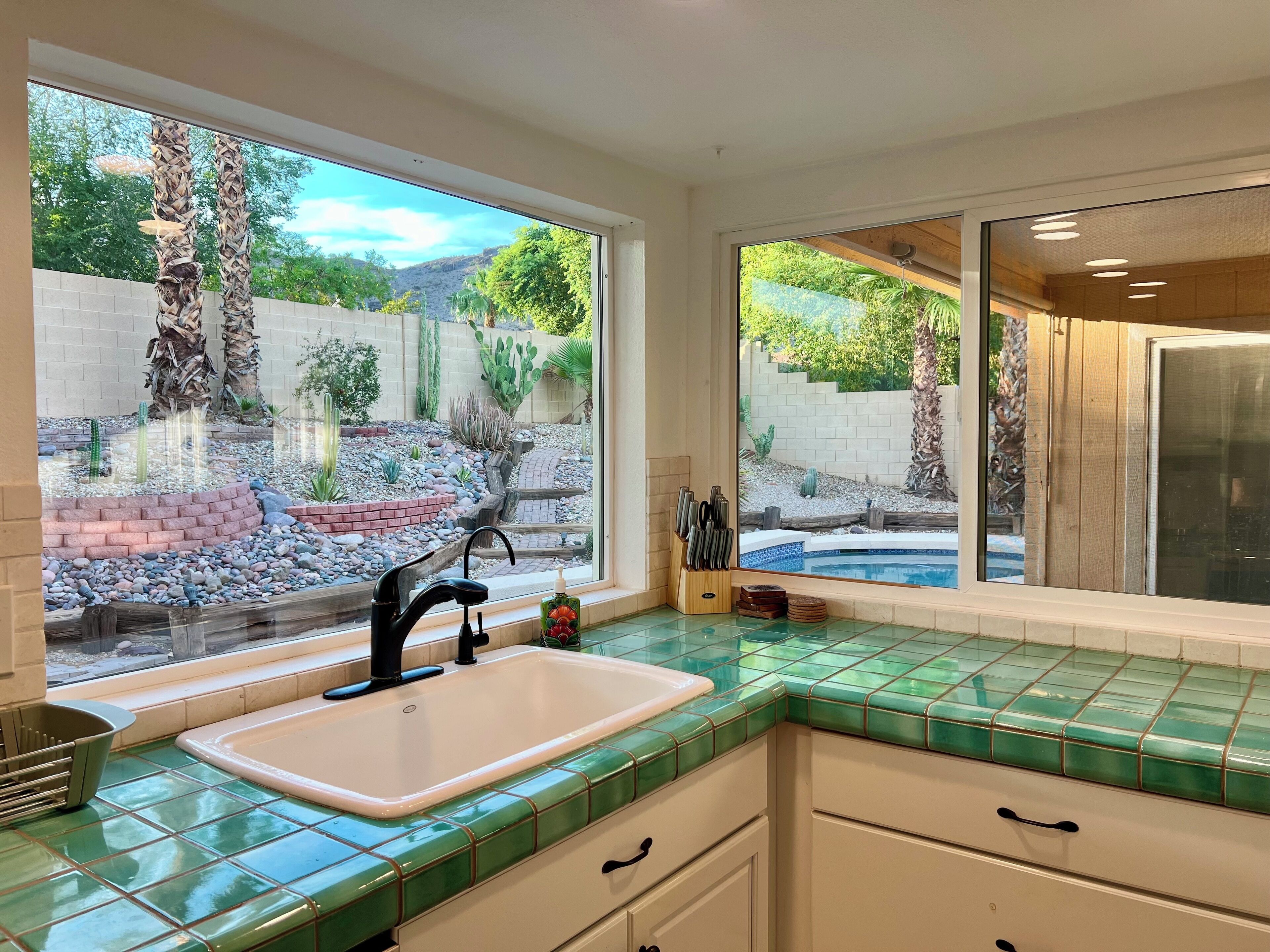 A kitchen with a large window, green tiled countertops, a white sink, and a view of a landscaped garden with cacti and a pool.