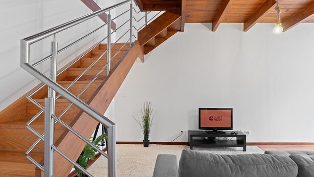 Un salon moderne avec un escalier en bois, un canapé gris, un téléviseur affichant le message « PRÊT POUR LES INVITÉS » et un luminaire suspendu.