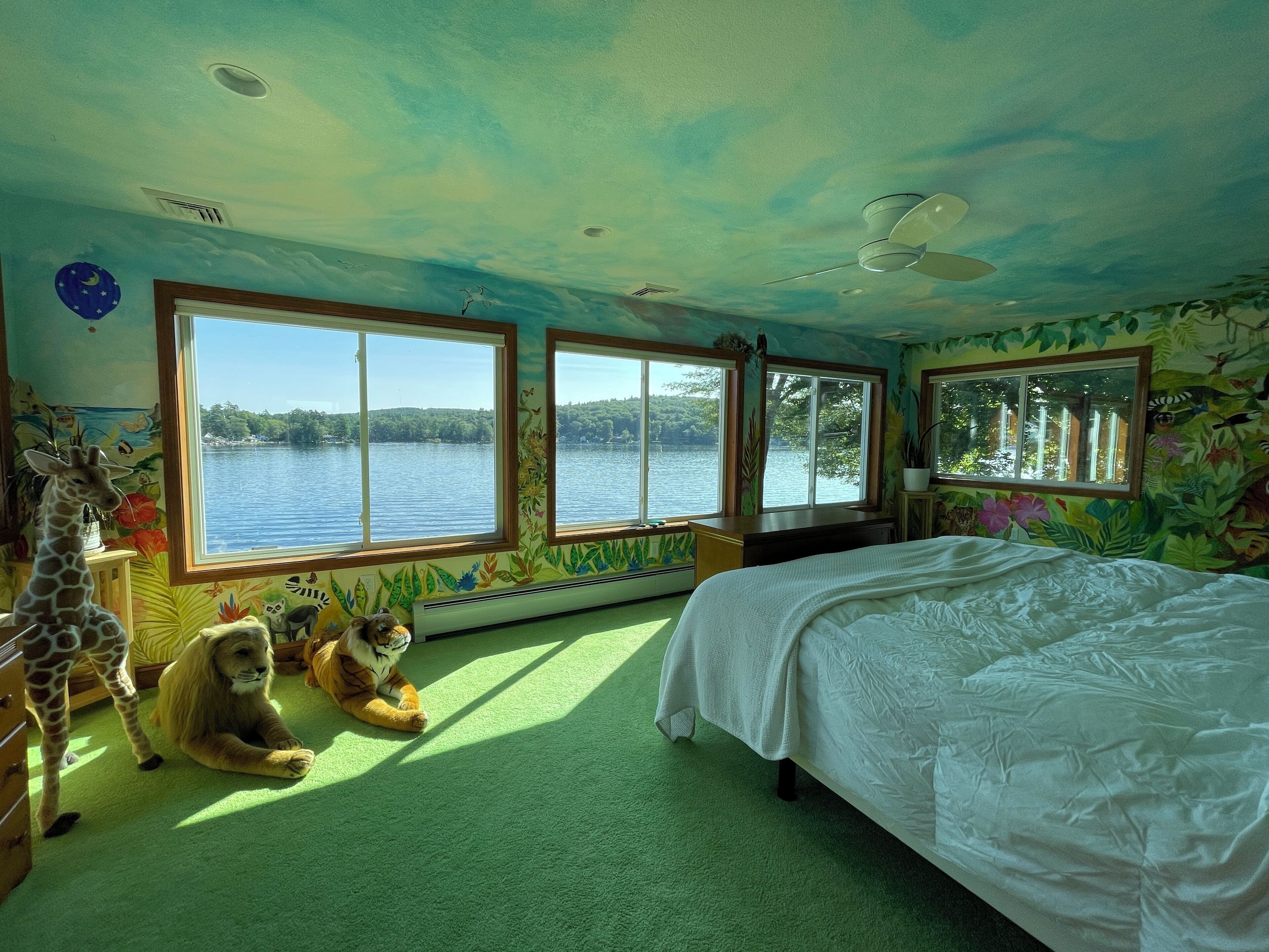 Quarto com cama, janelas amplas, estátuas de girafa e leão, e vista para um lago.