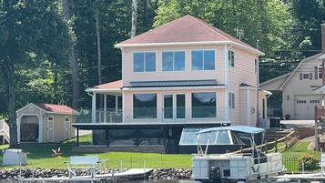 Una casa de dos pisos con un balcón grande, un muelle con un bote y un cobertizo pequeño en un entorno junto al lago.