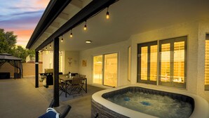 An outdoor patio area with a hot tub, seating, and a covered section with string lights. - Spacious 4 BR -SaltwaterHotTub -Pool -Sleeps 11 (Peoria)