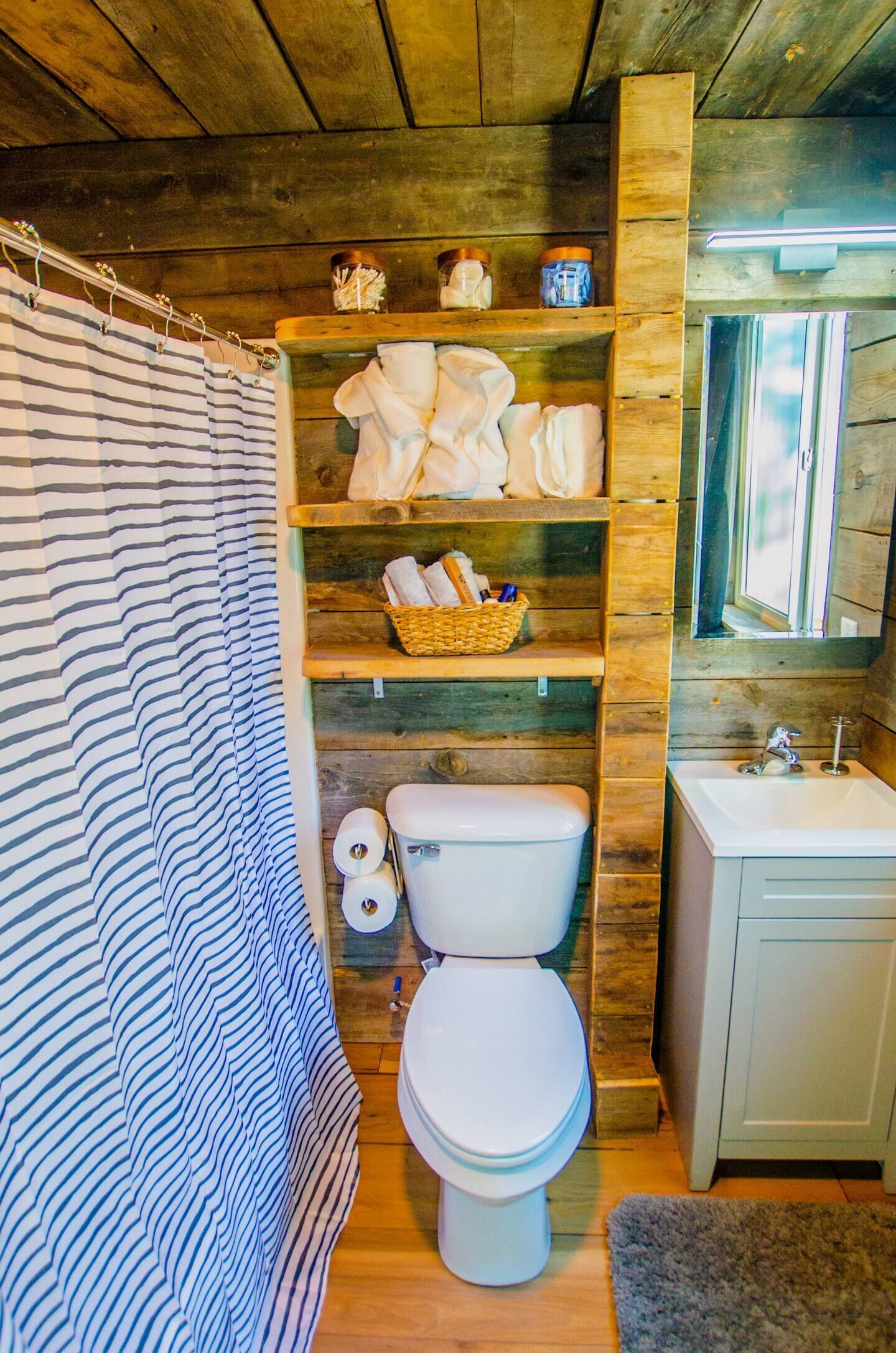 A bathroom with a striped shower curtain, wooden shelves with towels and toiletries, a white toilet, and a sink.