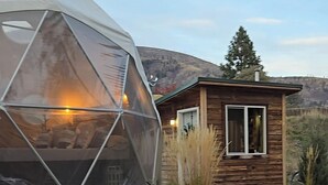 Una estructura de domo geodésico con luz cálida en su interior, junto a una cabaña de madera con ventana. Dos sillas Adirondack están ubicadas en un patio pavimentado.