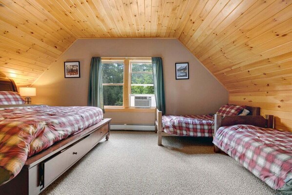 A room with a sloped ceiling, two beds with plaid bedding, a window with curtains, and a wall-mounted heater. - Classic Maine Chalet • Fireplace • Near Downtown (Bethel)