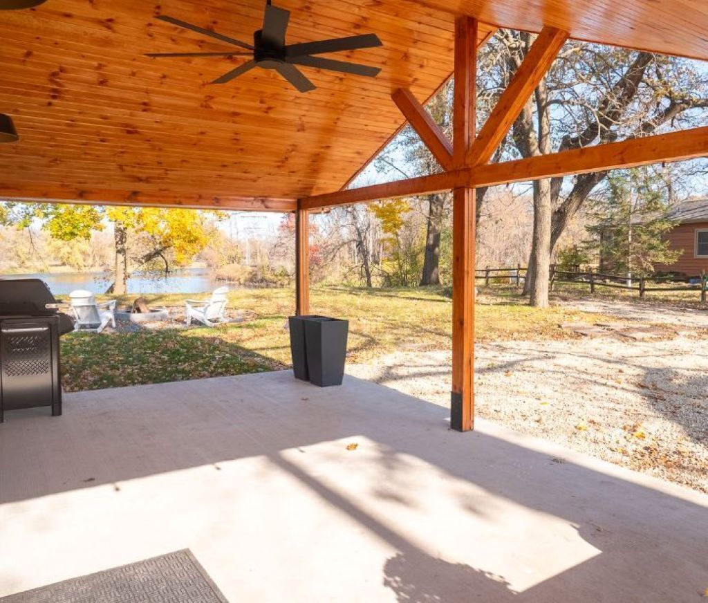 Une terrasse extérieure couverte, dotée d’un ventilateur de plafond, d’un sol en béton et offrant une vue sur un lac et des arbres en arrière-plan.