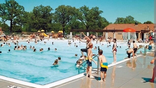 A swimming pool with multiple lanes, surrounded by trees and a covered area with umbrellas.