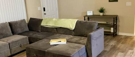 A living room with a sectional sofa, ottoman, and a small table with a plant.