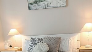 Una cama bien hecha con edredón blanco, dos mesitas de noche con lámparas, un cuadro colgado en la pared y suelo de baldosas.