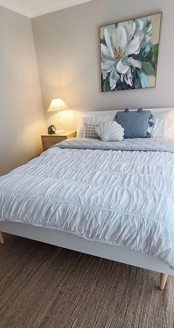 A neatly made bed with a white duvet and pillows, a bedside lamp, and a floral painting on the wall.
