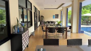 Un comedor moderno con vistas a la piscina, que incluye una mesa larga con sillas, una consola negra con objetos decorativos y un sofá de mimbre.