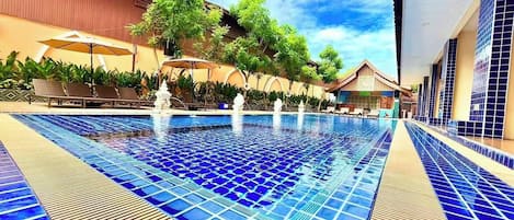Una piscina con azulejos azules, rodeada de sillas de descanso y sombrillas, con edificios y árboles al fondo.