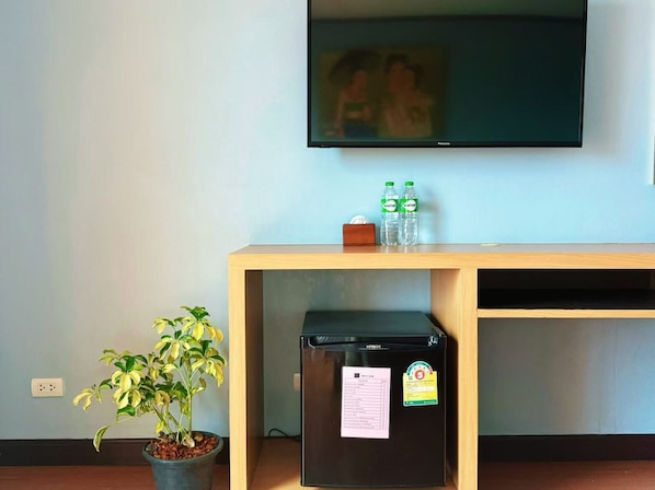 A room with a television mounted on the wall, a small refrigerator, a wooden console table with two bottles of water, and a potted plant.