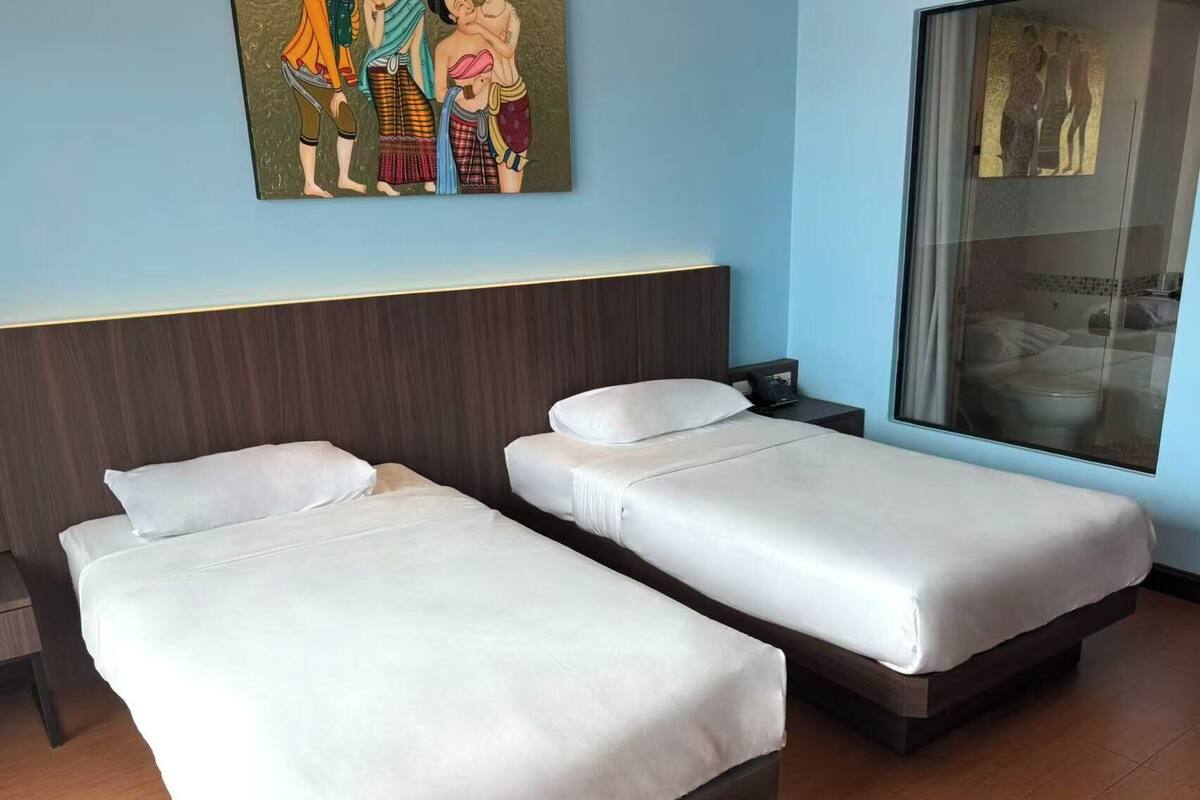 Habitación de hotel con dos camas individuales, cabecero de madera, mesita de noche, un cuadro en la pared y ventana con vista a un edificio.