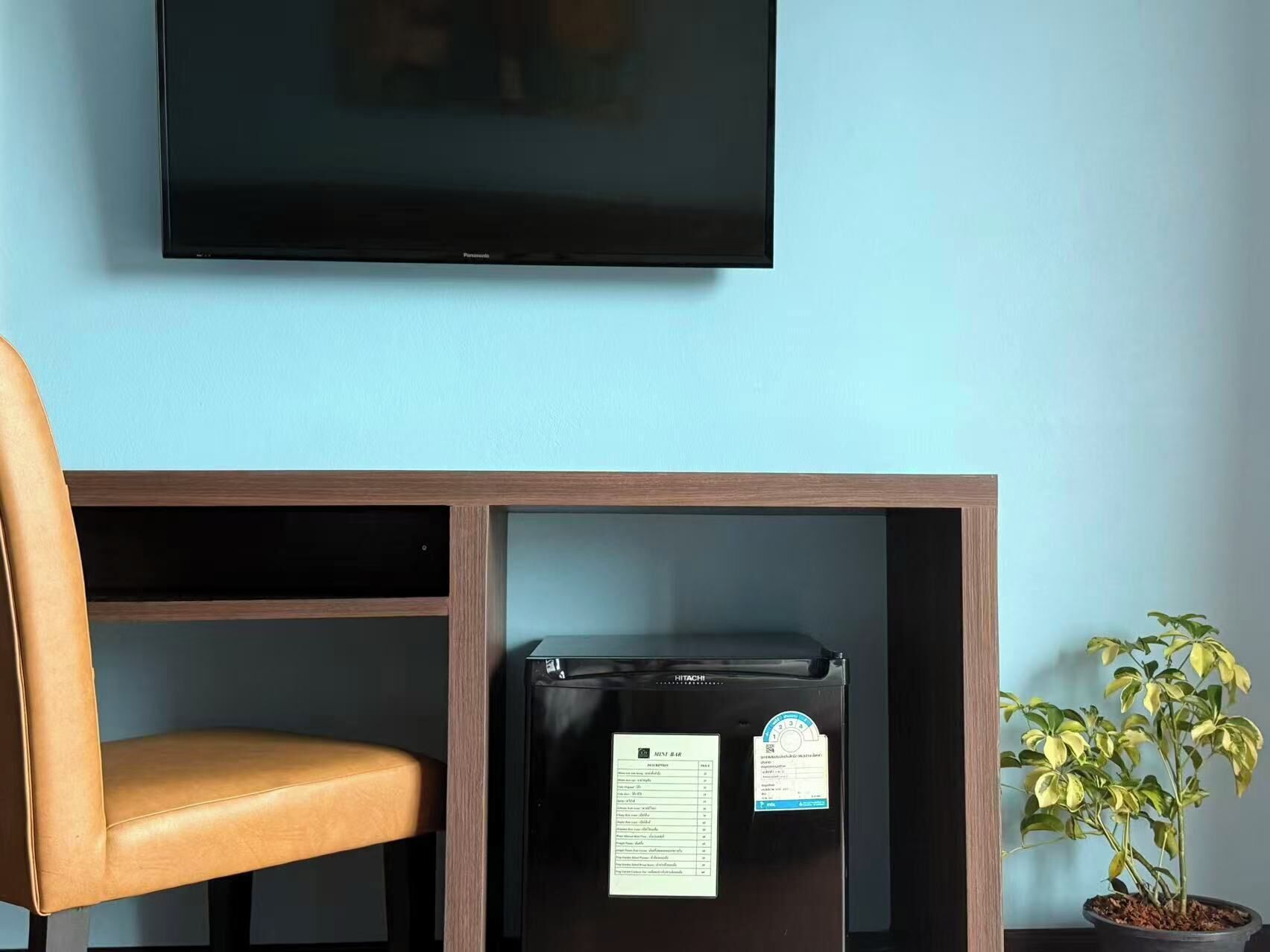 A room with a flat-screen TV mounted on the wall, a small black refrigerator, a leather chair, and a potted plant.