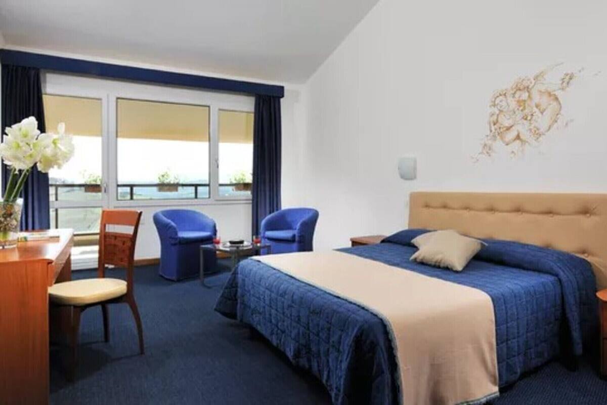 A hotel room with a large bed, a desk with a chair, two blue armchairs, and a view of the sea through large windows.