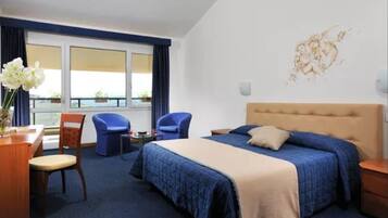 Camera d'albergo con un letto grande, una scrivania con una sedia, due poltrone blu e vista sul mare attraverso ampie finestre.