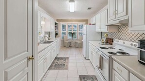 Une cuisine avec des meubles blancs, une cuisinière et un réfrigérateur. Il y a une fenêtre avec des stores et une porte qui donne sur une autre pièce.