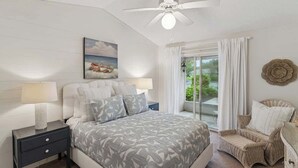 Une chambre à coucher comprenant un lit, des tables de chevet, un ventilateur de plafond, une fenêtre avec des rideaux et un fauteuil en osier.