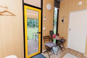 Ein Zimmer mit Holzboden, einer gelben Tür, einem kleinen Tisch mit Stuhl und Blick auf eine Terrasse und Bäume.