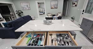 A modern kitchen with a marble countertop, a built-in drawer organizer with utensils, a dining area with a dark blue sofa, and a hallway with artwork. -   House in Tampa fl  , 3-bedroom , 2 bathroom  , pool (Tampa)
