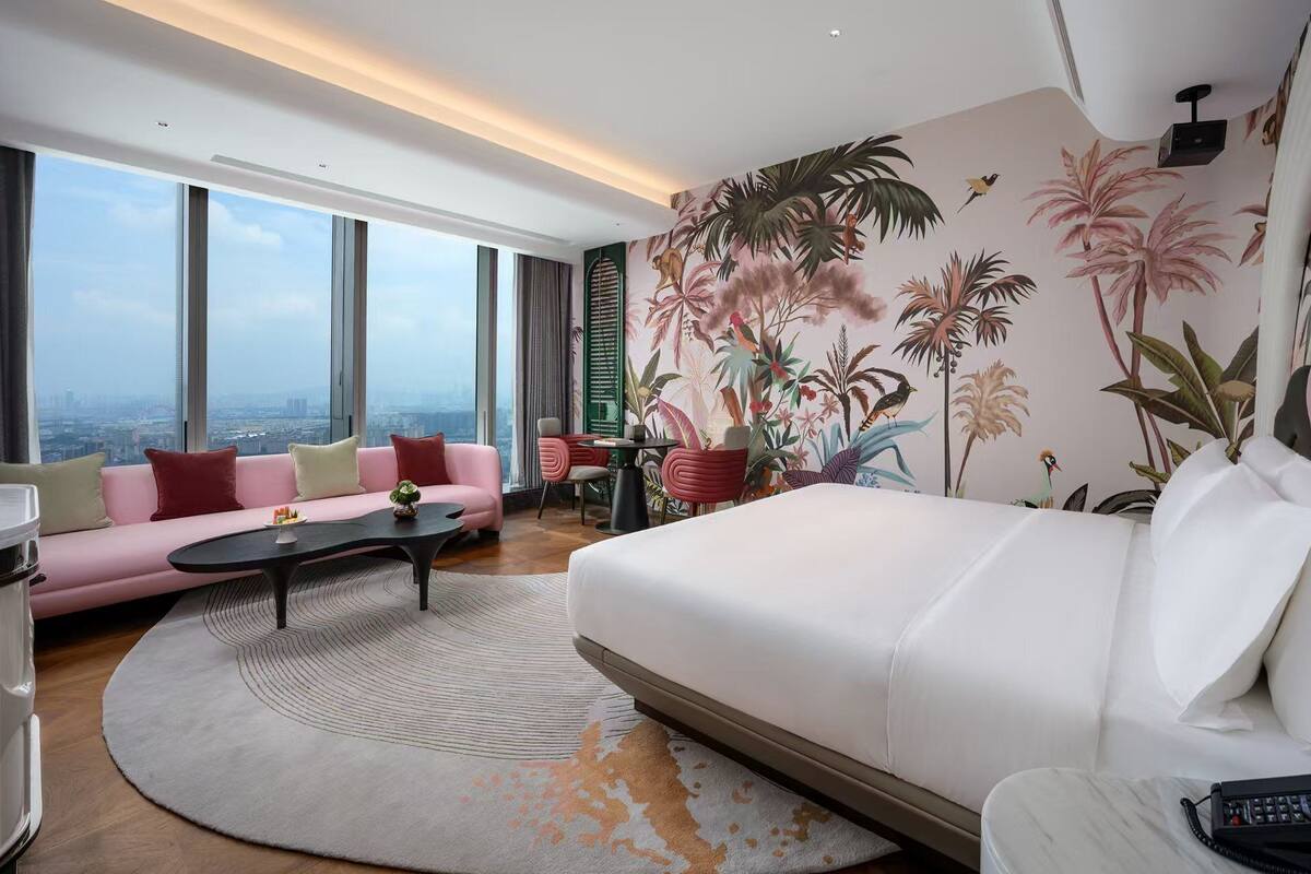 Ein modernes Hotelzimmer mit einem großen Bett, einem rosa Sofa, einem runden Couchtisch und Blick auf die Stadt durch große Fenster.