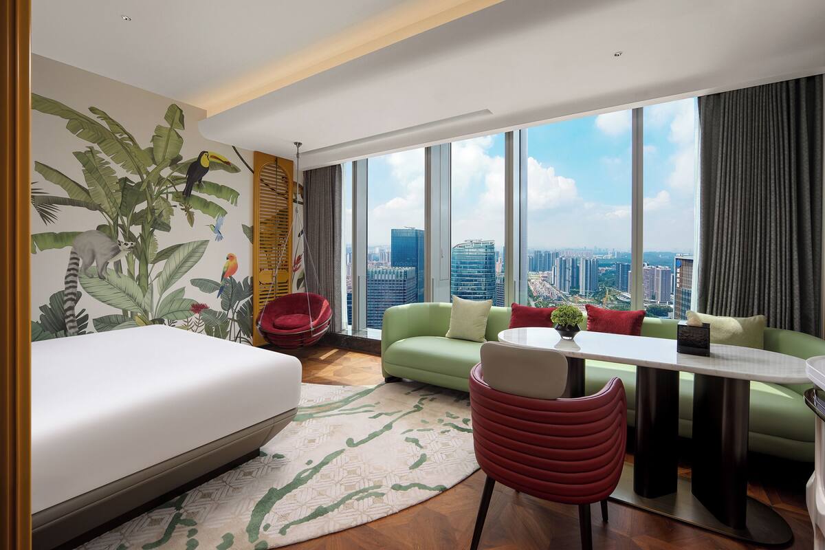Ein modernes Hotelzimmer mit einem Bett, einer grünen Couch, einem roten Sessel, einem Esstisch und einem großen Fenster mit Blick auf die Stadt.