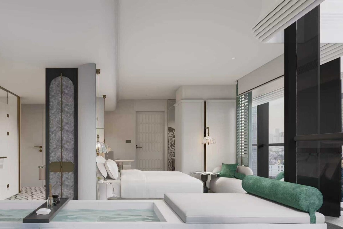 Una habitación de hotel moderna con una cama grande, una zona de estar con un sofá verde, una mesita y un ventanal con vistas a la ciudad.