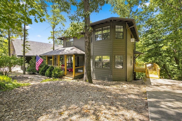 A two-story cabin with a porch and a deck, surrounded by trees and a gravel driveway.