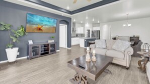 A spacious living room with a large painting, a ceiling fan, and a modern kitchen in the background. - Modern Spacious Family Retreat- Comfort & Style Await (El Paso)