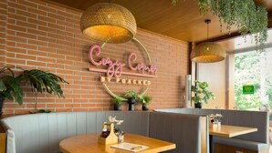 A cozy cafe with a neon sign, wooden tables, grey cushioned seating, and hanging plants.
