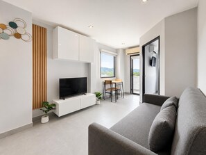A modern living room with a flat-screen TV, a sofa, a dining area with a table and chairs, and a large window with a view. - Boutique Bliss 1BR T1-506 (Chalong)
