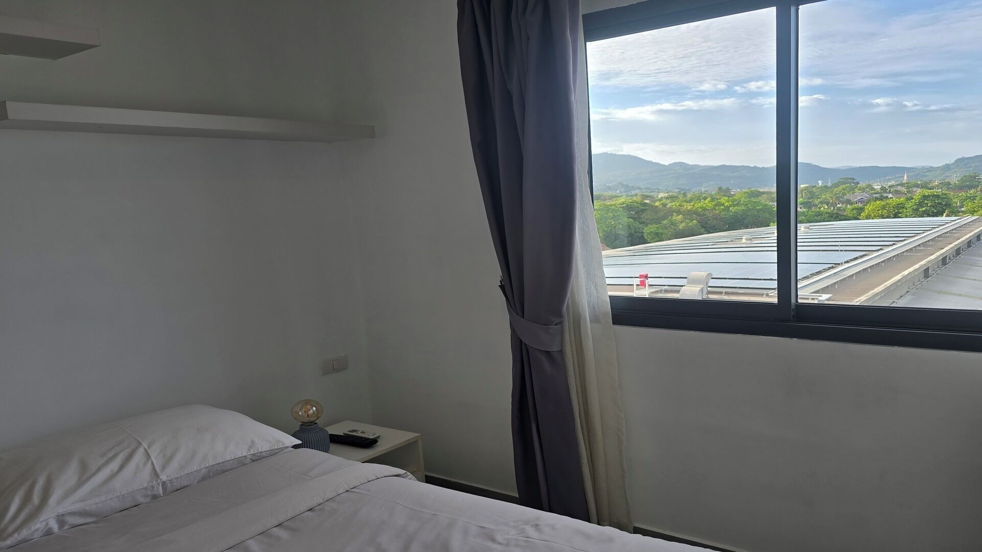 A hotel room with a bed, a nightstand, a lamp, and a window with a view of solar panels and a distant landscape.