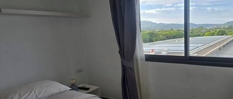 A hotel room with a bed, a nightstand, a lamp, and a window with a view of solar panels and a distant landscape.