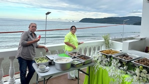 Un balcon avec une table dressée pour un repas, donnant sur une étendue d’eau, avec une côte et des montagnes visibles au loin.