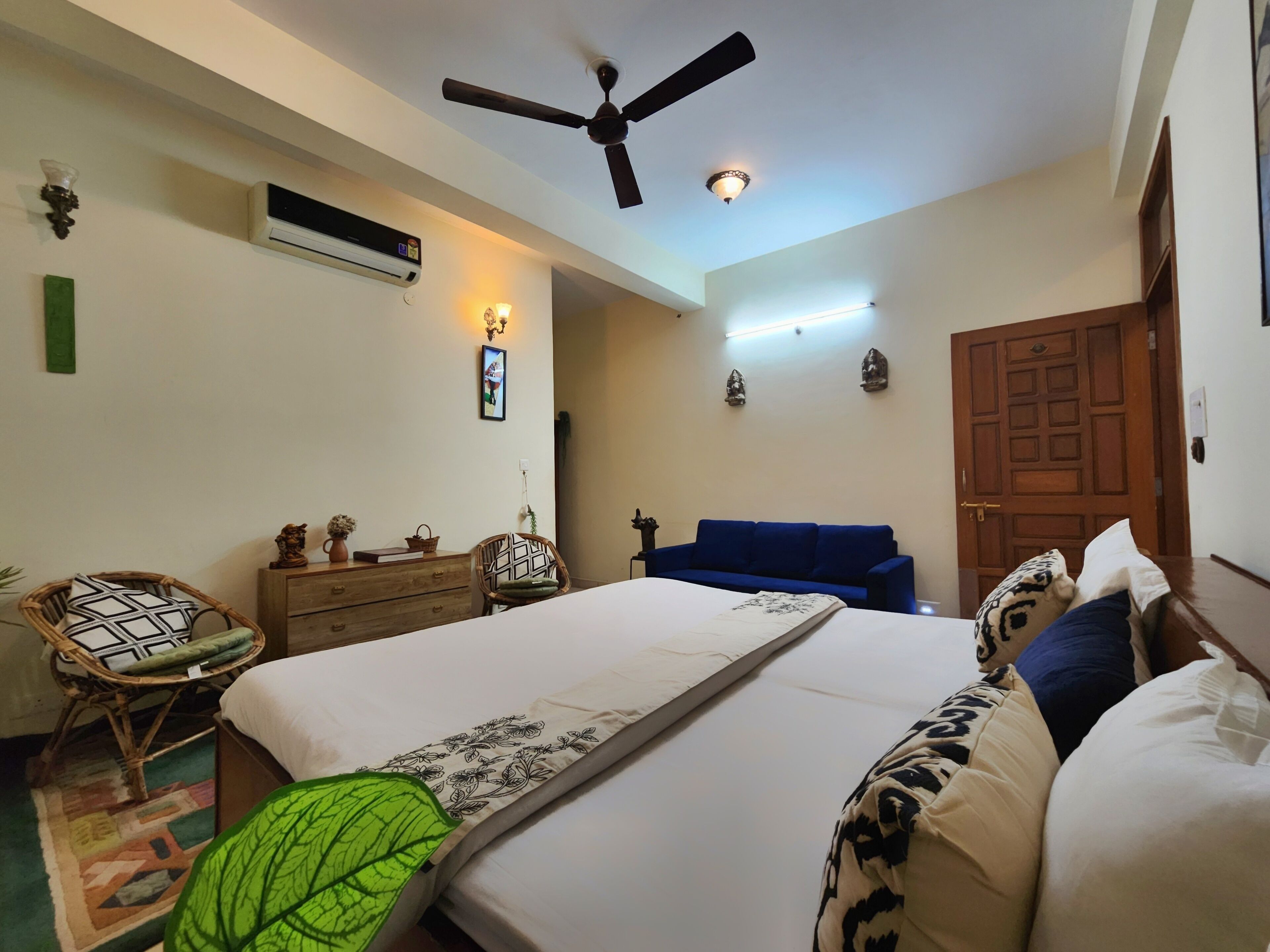 A bedroom with a bed, a blue sofa, a wooden wardrobe, a ceiling fan, and an air conditioning unit.