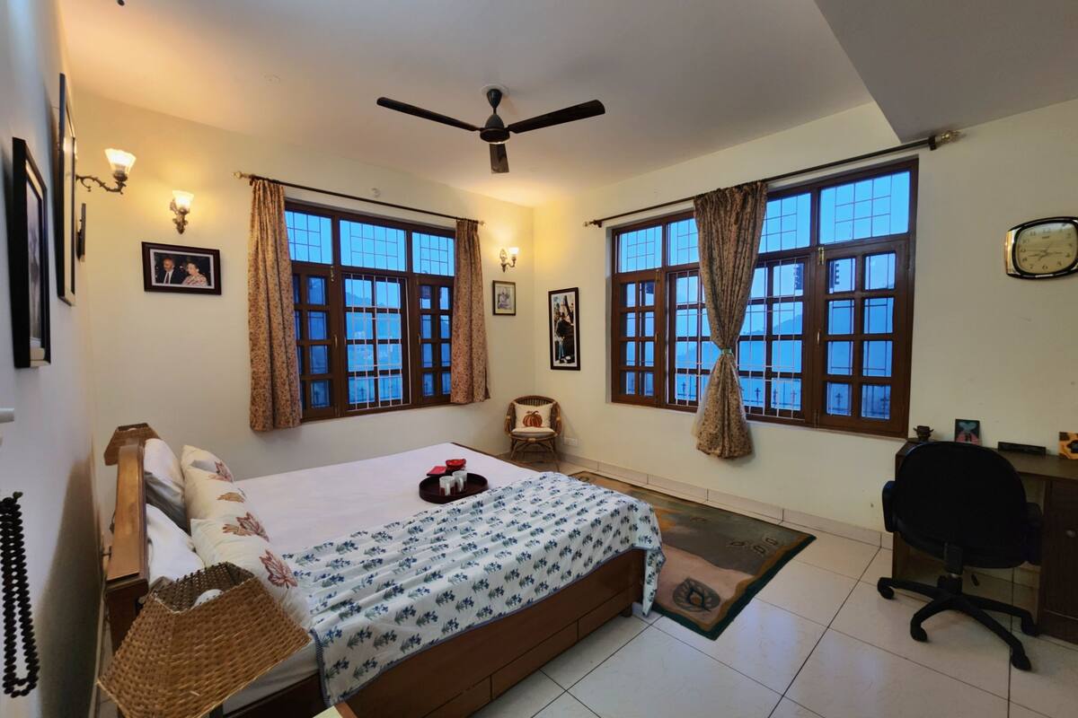 A bedroom with a bed, a desk, two windows with curtains, a ceiling fan, and a wall clock.
